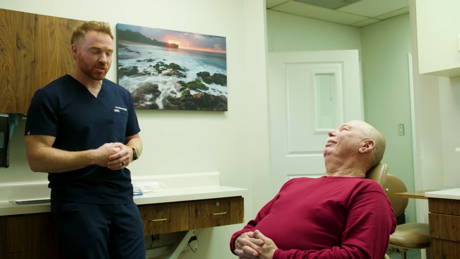 Dr. Tim Rauch standing in a consult room, explaining a treatment plan to a patient in the chair, with a coastal Hawaii sunset print behind them